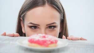 Beautiful girl is looking at unhealthy donut with appetite. It is situated on a table. Isolated on a white background